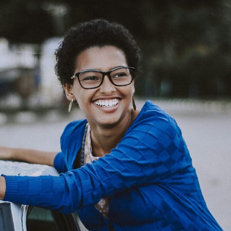 Smiling woman with short curly hair, wearing glasses and a blue cardigan, leaning on a car.